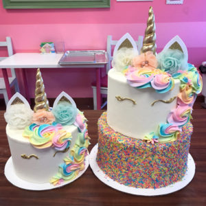 A two tier unicorn cake is pictured in a bakery. The bottom tier is completely covered in rainbow sprinkles and the top tier is a white unicorn. It has a hand painted gold fondant horn, white and gold fondant ears and a pastel rainbow mane.