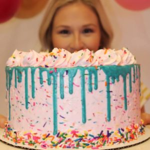 A blond woman is holding a light pink cake that has rainbow sprinkles mixed into the buttercream. there are rainbow sprinkles pressed onto the base of the cake and there is a teal chocolate drip around the top edge of the cake and on top of the drip is a buttercream rosette border. The rainbow sprinkles mimic a confetti celebration