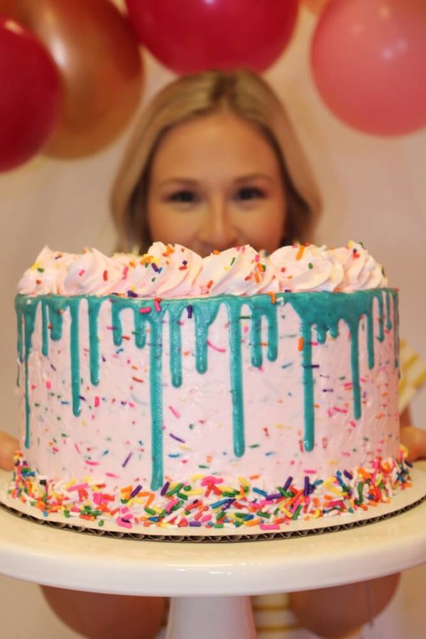 A blond woman is holding a light pink cake that has rainbow sprinkles mixed into the buttercream. there are rainbow sprinkles pressed onto the base of the cake and there is a teal chocolate drip around the top edge of the cake and on top of the drip is a buttercream rosette border. The rainbow sprinkles mimic a confetti celebration