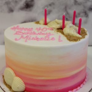 A buttercream cake with an ombre design of hot pink to light orange to white. Crushed graham crackers that resemble sand and white chocolate shells are in a semi-circle around the top outter edge of the cake.The colors and shell combination makes you feel like watching a sunset on the beach. The theme is she sells seashells by the seashore.