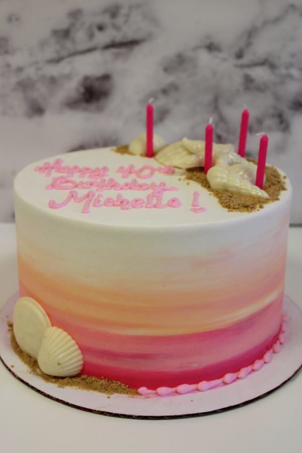 A buttercream cake with an ombre design of hot pink to light orange to white. Crushed graham crackers that resemble sand and white chocolate shells are in a semi-circle around the top outter edge of the cake.The colors and shell combination makes you feel like watching a sunset on the beach. The theme is she sells seashells by the seashore.