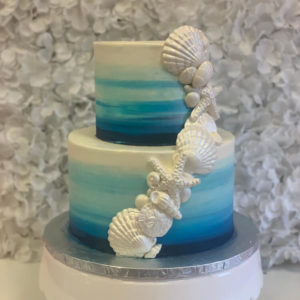 A two teir birthday cake with a sky blue ombre design. White chocolate shells with a pearl sheen cascade up and around the cake for a birthday by the beach celebration