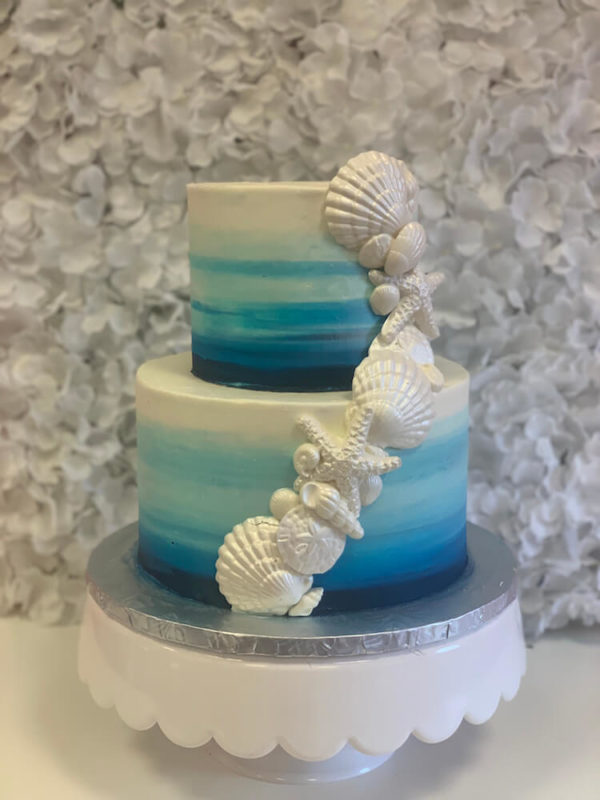 A two teir birthday cake with a sky blue ombre design. White chocolate shells with a pearl sheen cascade up and around the cake for a birthday by the beach celebration