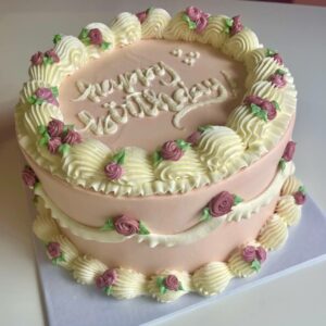 A mauve, round cake with a vintage birthday cake design. There is white buttercream pipping detail and dark pink buttercream roses. The top of the cake reads "happy birthday!"