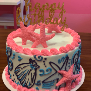 A white buttercream cake inspired by Lily Pulitzer. This beach themed cake has a navy blue and sky blue shell and starfish design around the cake. Hot pink shell piping is around the boarder with three hot pink chocolate starfish on top of the cake. There is a gold glittery happy birthday cake topper and the cake sits on a wooden table.