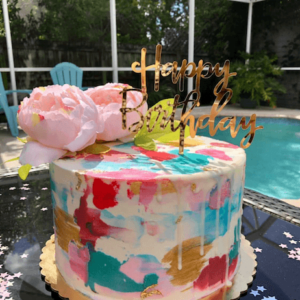 An abstract decorated cake with pink, teal, salmon and white buttercream sits atop a table beside a backyard pool