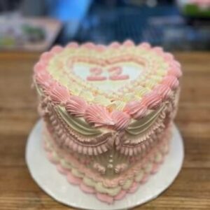 A heart shaped cake with white and light pink buttercream. The cake is decorated in the style of vintage wedding cakes, with large shell piping, and delicate buttercream ruffles.